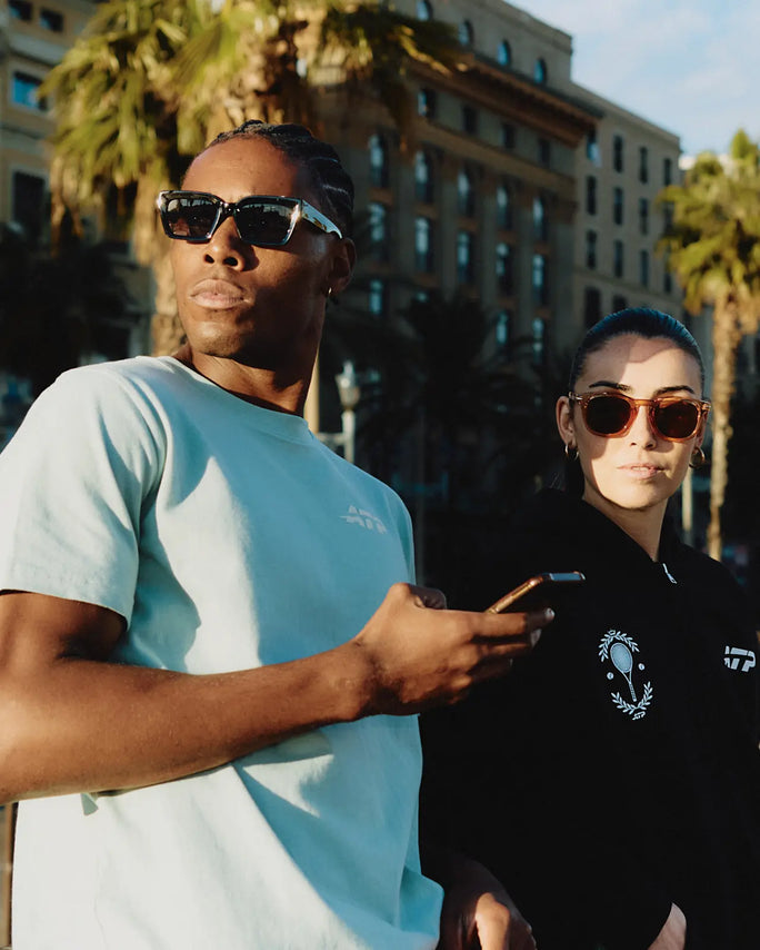 Two people wearing ATP clothing in an urban setting with palm trees and a building in the background.