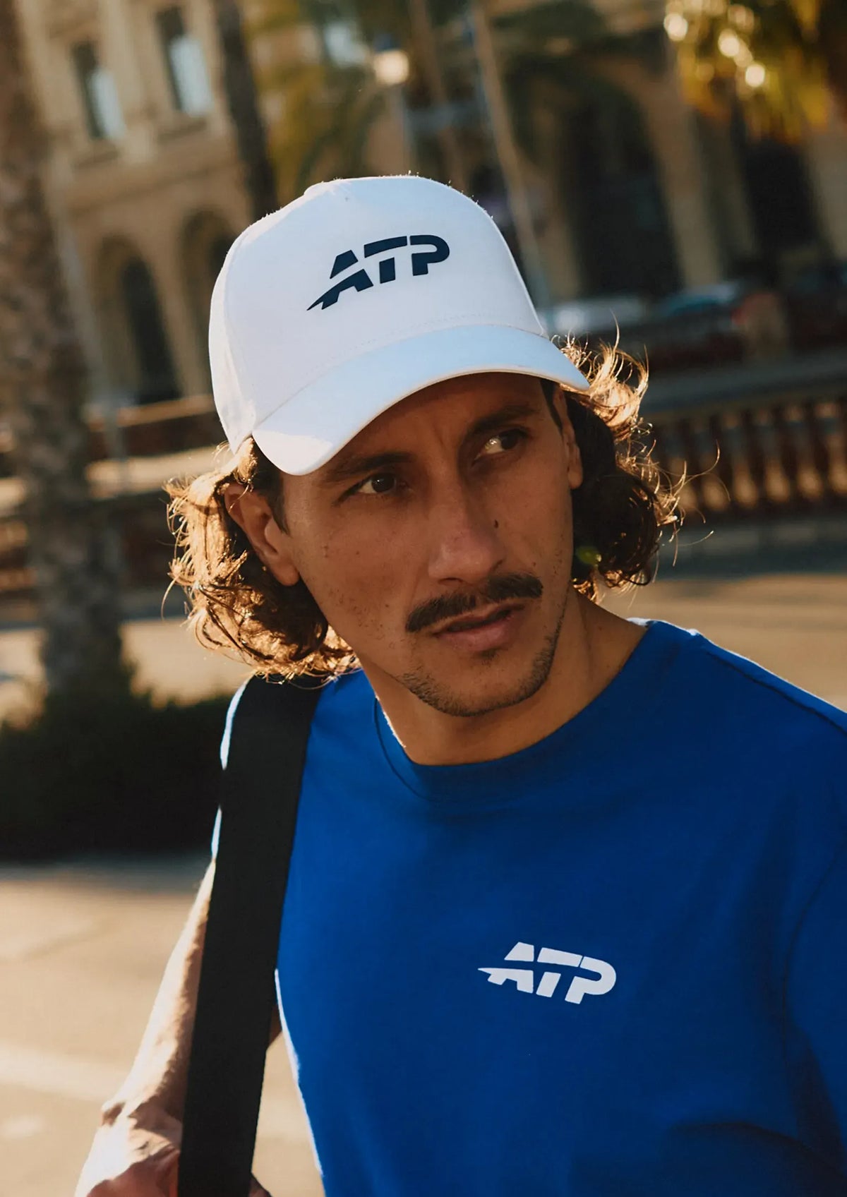 Man wearing a white cap and Blue T-Shirt with 'ATP' logo on a street with palm trees and buildings in the background.