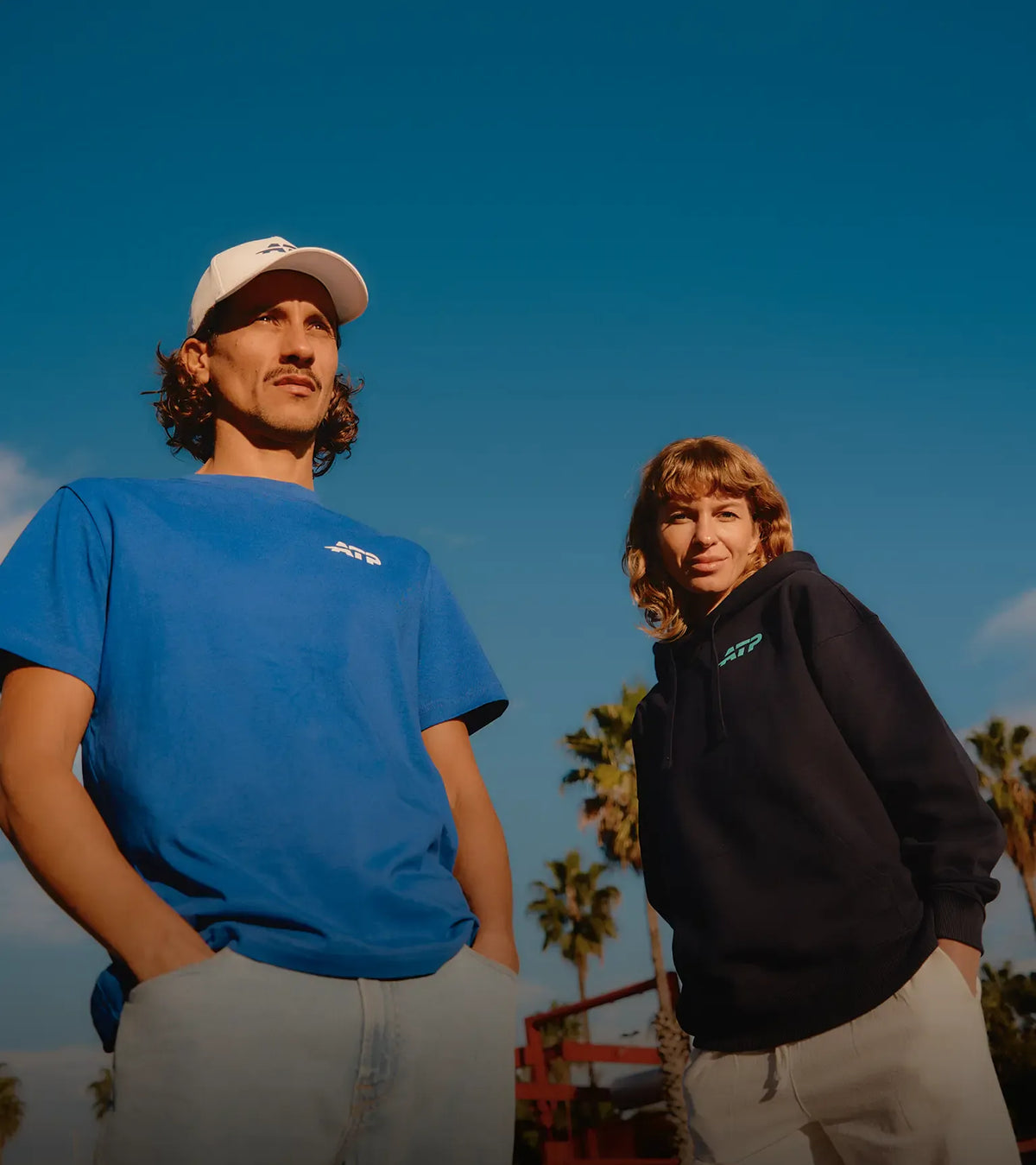 Two people standing outdoors with palm trees and a clear blue sky in the background