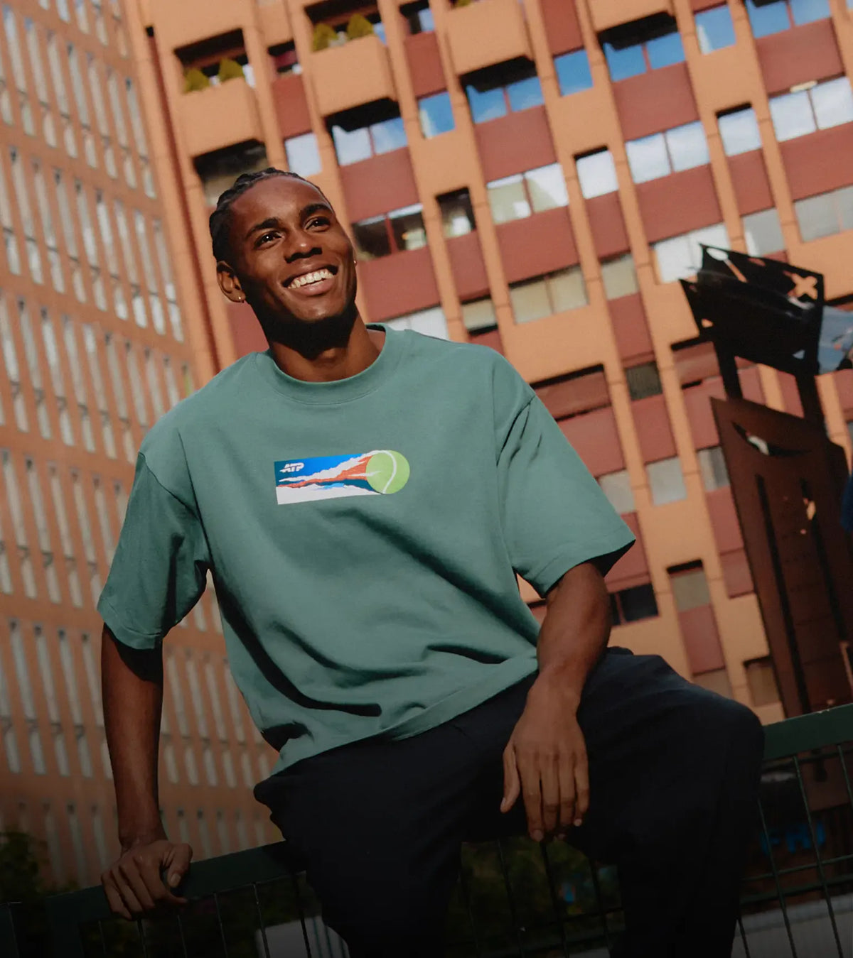 Man wearing a green t-shirt with a logo, sitting on a ledge with a building in the background