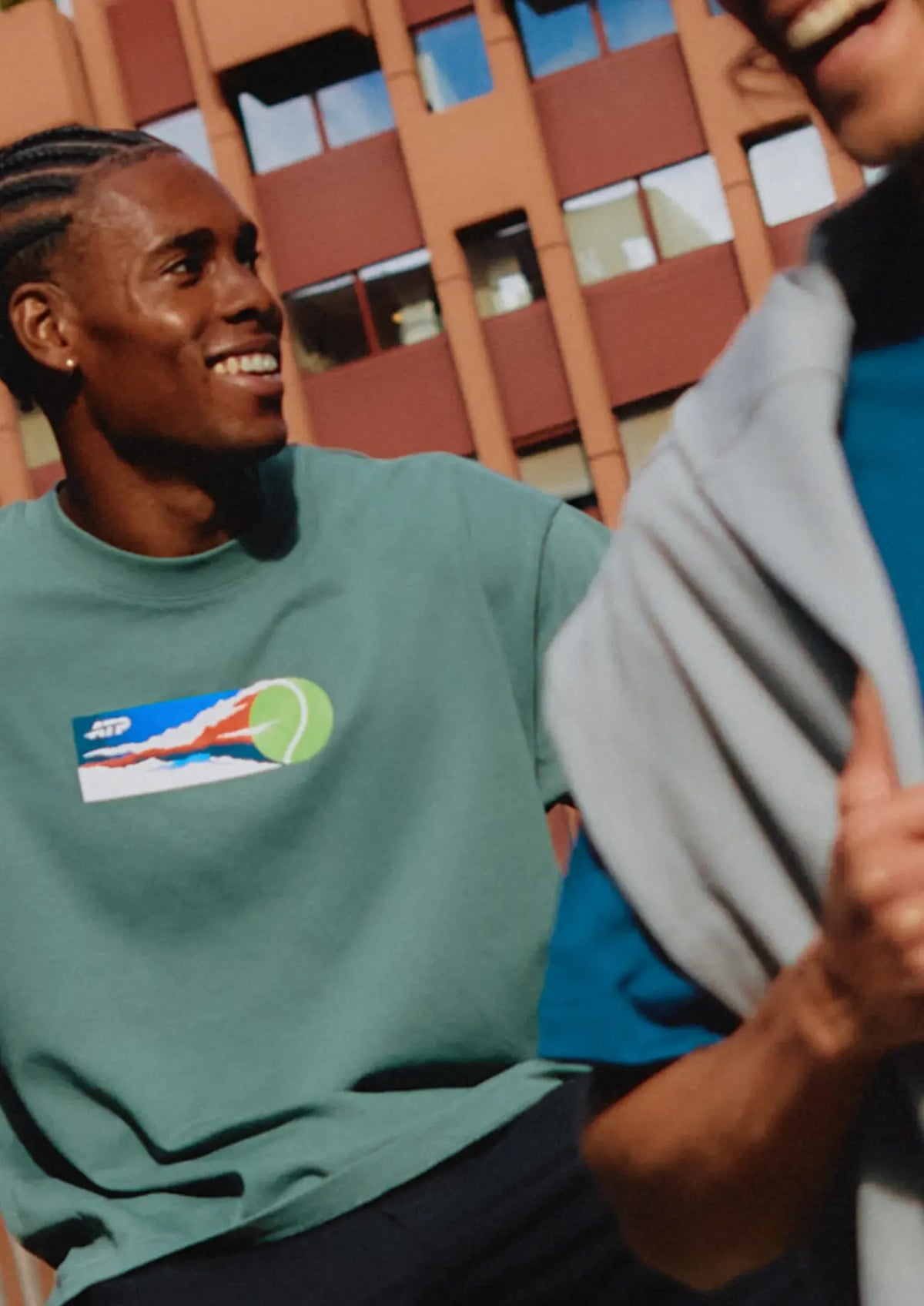 Two people wearing green and blue t-shirts with tennis logos, standing outdoors in front of buildings.