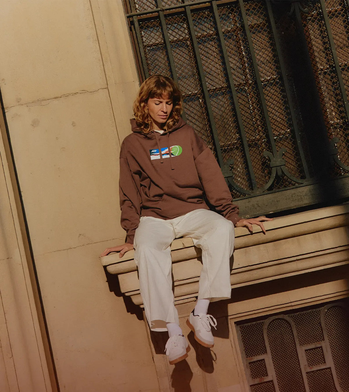 Woman wearing a brown sweatshirt with tennis graphic sitting against a wall.