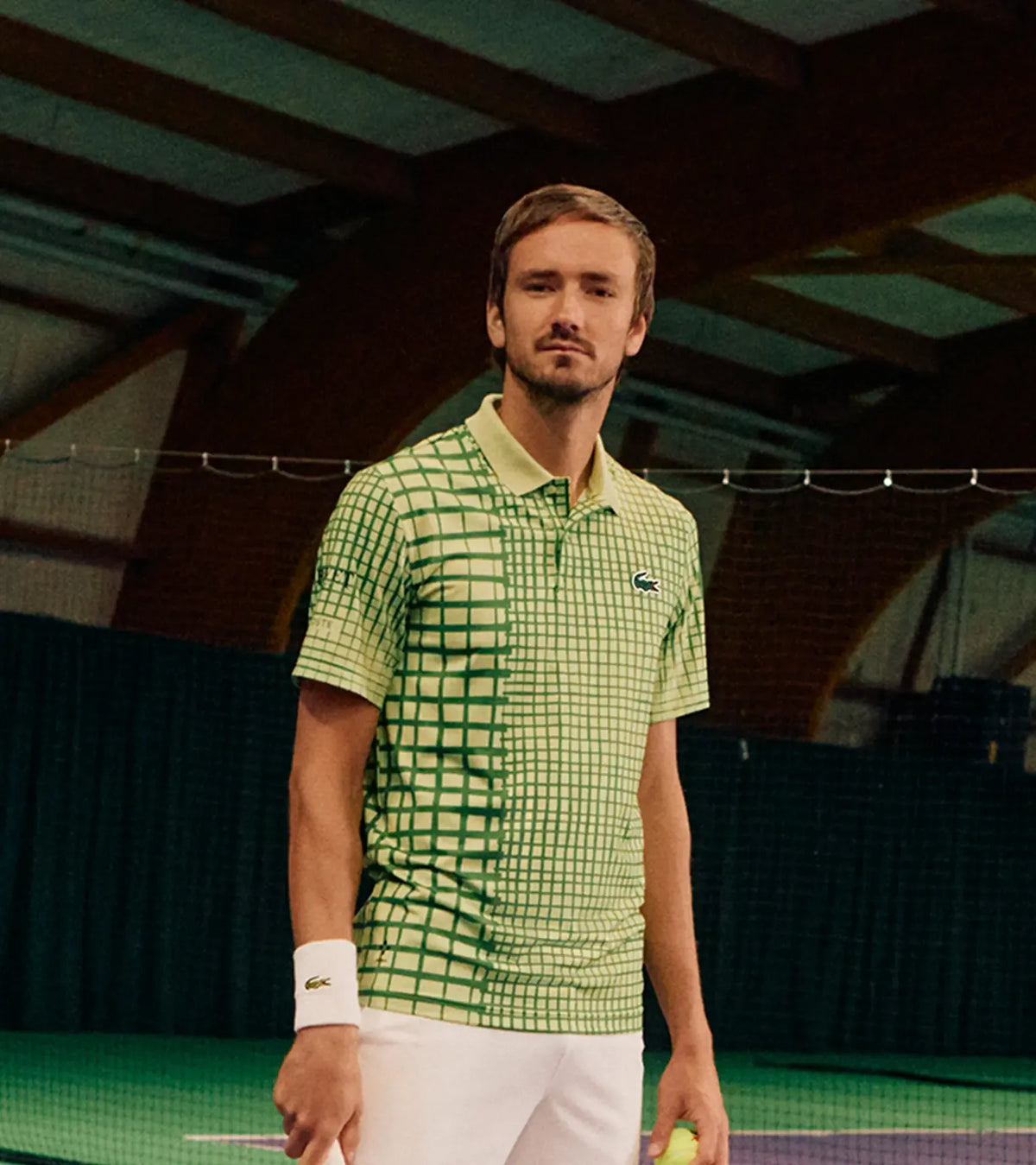 Daniil Medvedev in a green Lacoste T-shirt standing on a tennis court inside a large indoor sports facility.